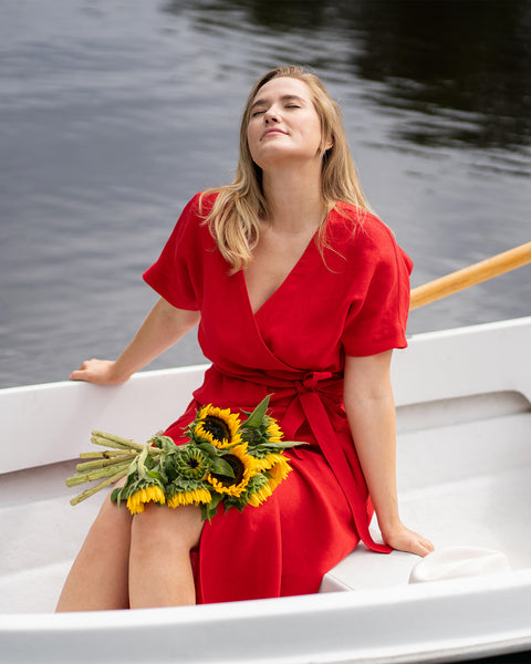 A woman relaxes in a small white rowboat on calm water, tilting her head back with her eyes closed to peacefully soak in the sunlight. She is wearing a bright red linen wrap dress with a vibrant bouquet of fresh sunflowers resting comfortably on her lap.