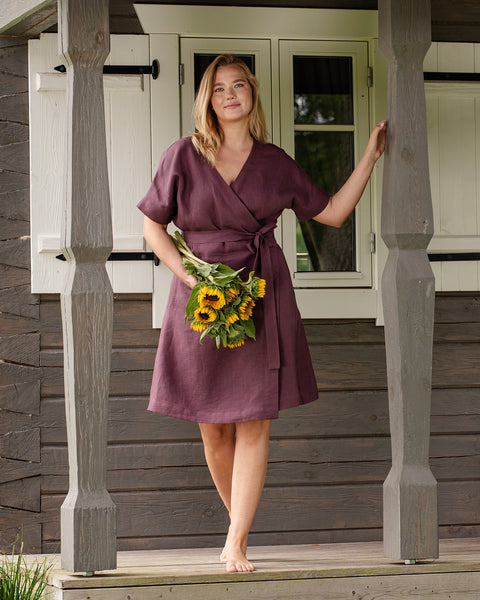 A woman stands barefoot on a wooden porch between two carved pillars, smiling softly at the camera. She is dressed in a shadow purple linen wrap dress with a tied waist, carrying a bunch of bright sunflowers in one hand.