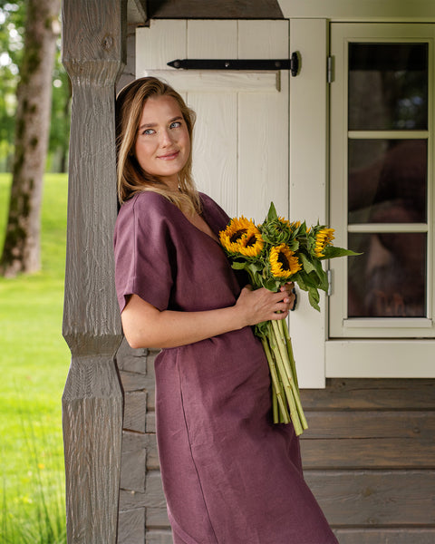 A woman stands gently leaning against a wooden pillar on a porch, looking thoughtfully toward the sunlit green yard. She is wearing a shadow purple, short-sleeve linen wrap dress and holding a bright bouquet of sunflowers.