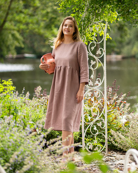 A barefoot woman stands near a white decorative garden archway, wearing a long-sleeve, faded rose linen dress. She smiles softly at the camera while holding a decorative terracotta pitcher comfortably against her side.