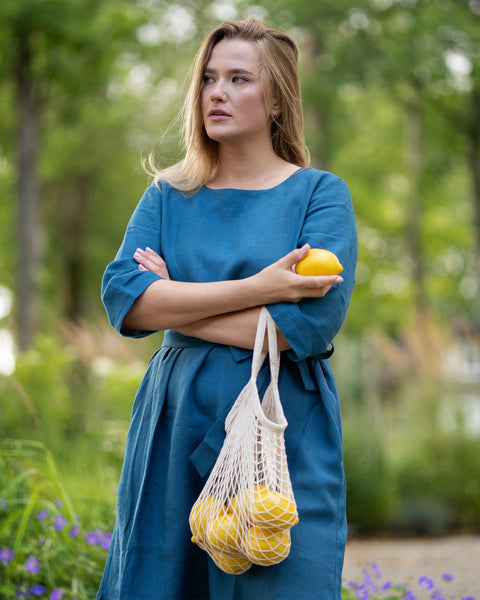 A woman with crossed arms gazes thoughtfully to the side while standing in a lush green garden. She wears a cobalt blue linen dress with a tie waist, holding a fresh lemon with a string bag of lemons hanging from her wrist.