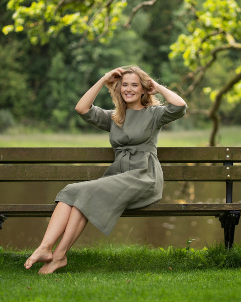 A woman sits on a wooden bench in a park wearing a green dress with her legs elegantly crossed. She holds both hands up near her face while looking slightly upward into the distance.