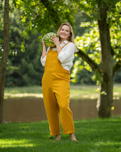 A cheerful, barefoot woman stands on the grass near a serene lake, playfully holding a small whole watermelon up to her shoulder. Her summery outfit features a pure white collared linen shirt worn underneath a bright, spicy yellow linen jumpsuit.