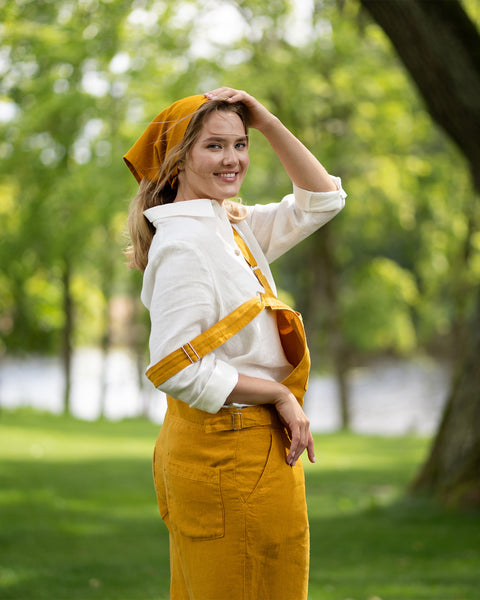 A woman stands in a sunlit green park, glancing happily over her shoulder directly at the camera with one hand resting gently on her head. She is dressed comfortably in a pure white linen shirt paired with a spicy yellow linen jumpsuit and a matching yellow bandana.