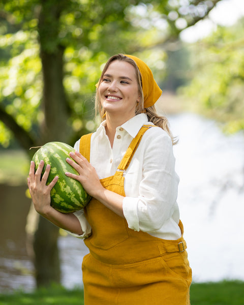 A cheerful woman stands near a tranquil lake, cradling a whole watermelon in her arms and smiling thoughtfully into the distance. She is styled in a pure white linen shirt layered under a spicy yellow jumpsuit with a coordinating bandana.