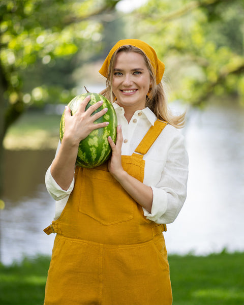A smiling woman stands outdoors in a sunny park, affectionately hugging a whole watermelon to her chest. She is wearing a pure white linen button-down shirt layered under a spicy yellow jumpsuit, completed with a matching yellow bandana.