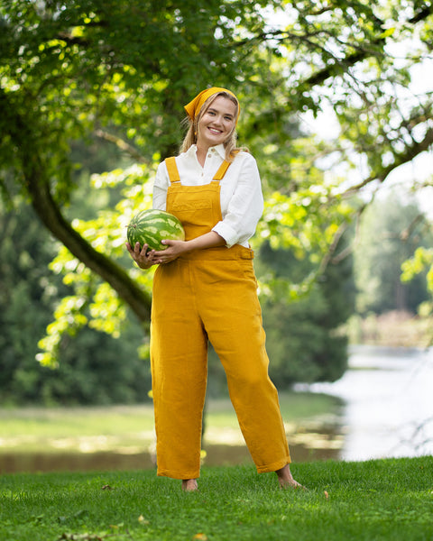 A smiling, barefoot woman stands on lush green grass, holding a whole watermelon at her waist. She is dressed in a vibrant spicy yellow linen jumpsuit over a pure white shirt, accessorized with a matching yellow bandana.