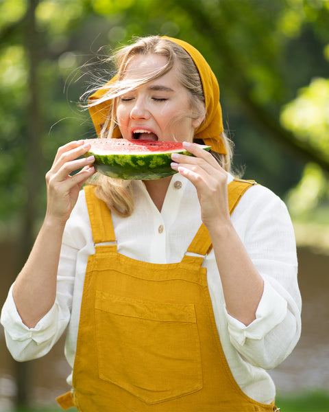 A woman stands outdoors with her eyes closed as she takes a joyful bite out of a large slice of fresh watermelon. She is wearing a spicy yellow linen jumpsuit paired with a crisp white shirt and a matching yellow bandana.