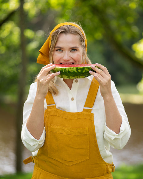 A woman smiles brightly at the camera while holding a juicy slice of watermelon up near her face. She is dressed for a sunny day in a pure white linen shirt layered under a spicy yellow jumpsuit and a matching head bandana.