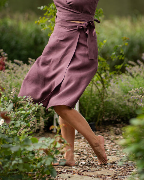 A barefoot woman stands playfully on a stone garden path, lifting one foot slightly off the ground. She is wearing a shadow purple linen wrap skirt, paired with a matching crop top that ties at the waist, surrounded by lush green foliage.