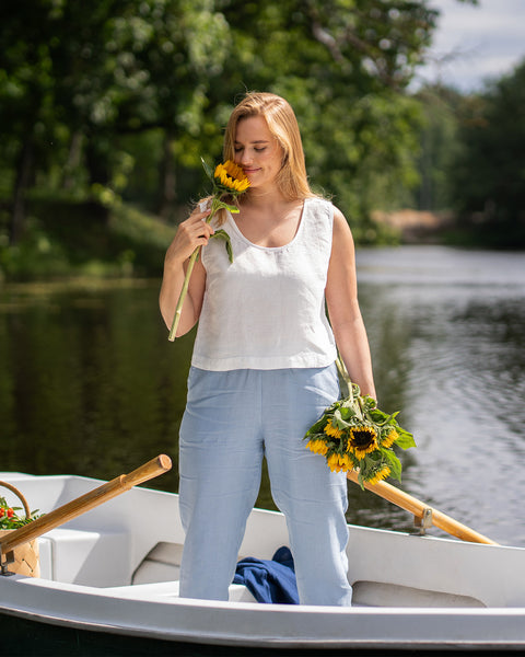 A blonde woman stands in a small rowboat on a serene river while holding a small yellow flower near her face. She is dressed in a white sleeveless top and light blue pants, with wooden oars resting across the boat.