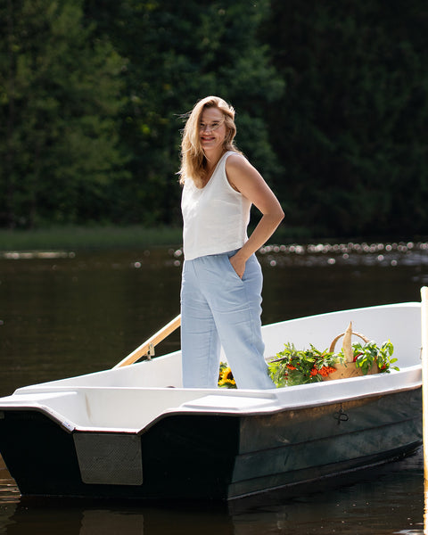 A smiling woman stands in a small white rowboat on a calm lake, wearing a pure white sleeveless linen top and light blue pants. A woven basket filled with fresh produce and bright sunflowers rests beside her in the boat.