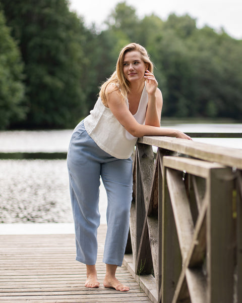 A blonde woman stands barefoot on a wooden dock wearing a sleeveless white linen top and relaxed light blue pants. She leans casually against a wooden railing, smiling thoughtfully with a serene lake and trees behind her.