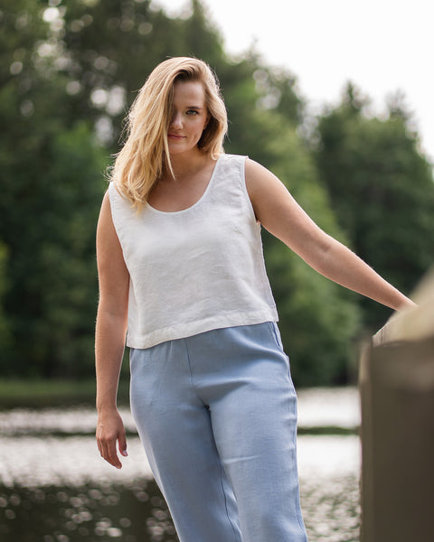 A woman stands on a wooden pier beside a lake, casually resting one hand on the wooden railing. She is wearing a pure white sleeveless linen top and light blue trousers against a lush backdrop of green trees.