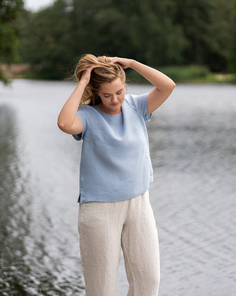 A woman stands near a tranquil lake, gently running her hands through her hair while looking downwards. Her comfortable outfit consists of a short-sleeve, cloudy blue linen top and loose-fitting, natural-colored linen pants.