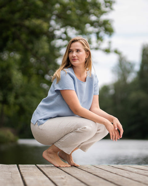 A barefoot woman crouches gracefully on a wooden pier by a lake, looking thoughtfully off into the distance. She is dressed in a short-sleeve, cloudy blue linen top and relaxed, natural-colored linen pants.