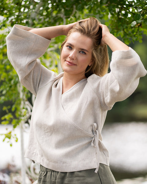 A woman stands outdoors near a body of water, smiling softly while holding her hair up with both hands. She is wearing a natural beige, long-sleeved linen wrap top with a side tie, paired with dark olive green pants.