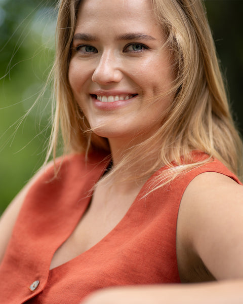 A close-up portrait shows a smiling woman wearing a sleeveless cinnamon red linen vest featuring a button detail. Her hair blows gently across her face in the breeze against a blurred backdrop of green foliage.