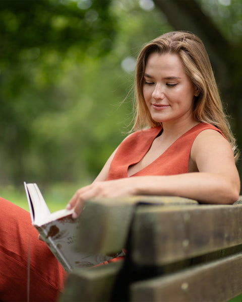 A woman sits on a wooden park bench, looking down and deeply engrossed in reading a book. She is dressed in a sleeveless cinnamon red linen outfit, set against a peaceful green park environment.