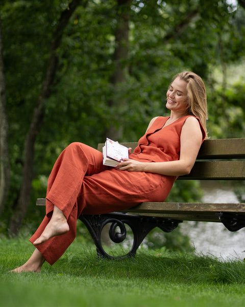 A woman wearing a cinnamon red linen vest and trousers sits on a park bench, resting a closed book in her lap. She smiles softly with her eyes closed, soaking in the serene, leafy outdoor surroundings.