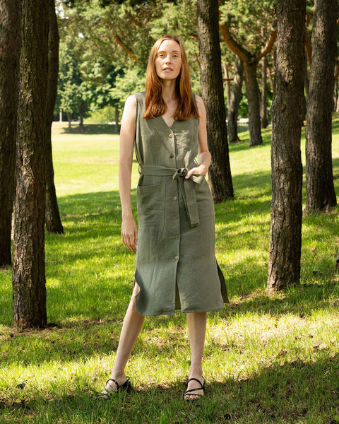 A fair-skinned woman with long, reddish-brown hair stands facing forward in a forest setting. She is wearing a sleeveless, stone green linen dress that falls to mid-calf. The dress has a V-neck and a row of buttons down the front, and a tie belt of the same fabric is tied around her waist. She is wearing dark, flat sandals. The background shows a grassy area surrounded by tall trees.