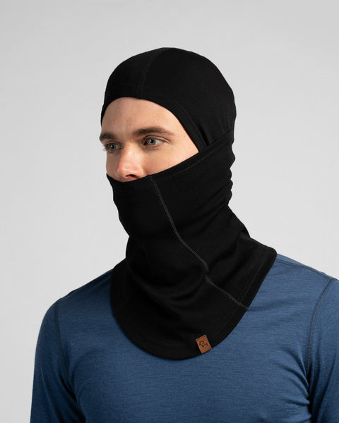 A studio portrait of a man looking off to the left side of the frame. He is wearing a black balaclava that covers his head and neck, but the front panel is pulled down under his nose, leaving his eyes and nose exposed. He is wearing a blue long-sleeved shirt against a plain light grey background.