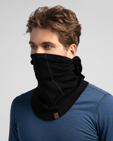 A studio portrait of a man with wavy brown hair looking off to the left. He is wearing a black balaclava pulled down off his head so it functions as a thick neck gaiter, covering his neck and lower face up to his nose. He is wearing a blue long-sleeved shirt against a plain light grey background.
