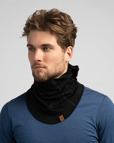 A studio portrait of a man with wavy brown hair and a short beard looking off to the left. He is wearing a black balaclava pulled completely down around his neck, functioning as a bunched neck warmer. He is wearing a blue long-sleeved shirt against a plain light grey background.