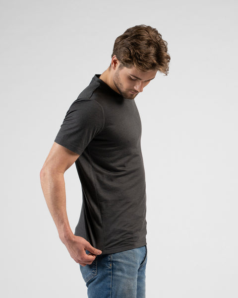 A side profile portrait of a man looking down while slightly adjusting the bottom hem of his charcoal grey short-sleeved t-shirt. He is wearing blue jeans against a light grey background.