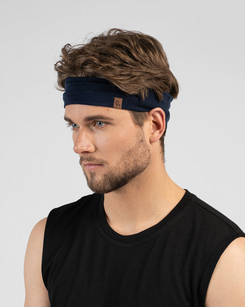 A studio portrait of a man looking down and to the left against a plain light grey background. He has wavy brown hair held back by a navy blue headband with a small logo tag, and is wearing a black tank top.