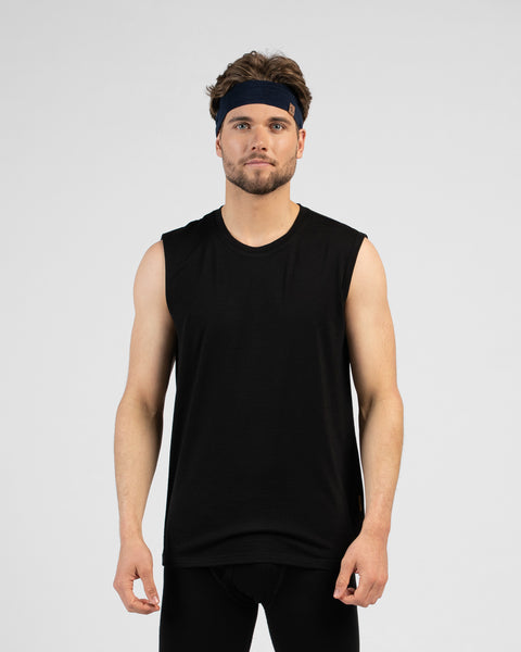 A studio portrait of a man standing and looking directly at the camera with his arms relaxed by his sides. He is wearing a black tank top, black pants, and a navy blue headband against a plain light grey background.
