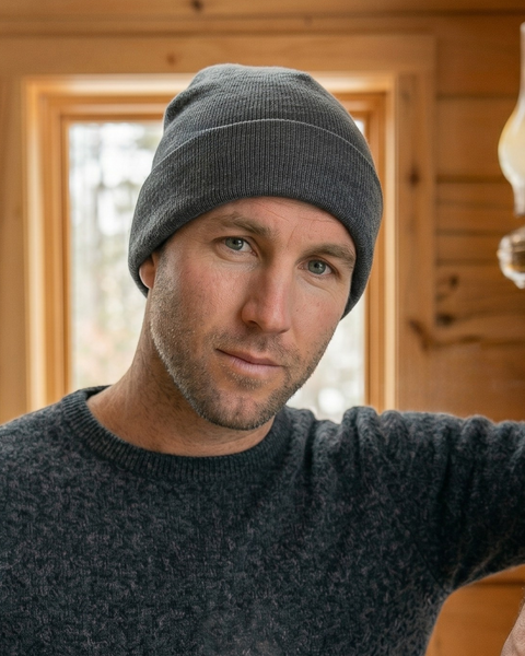 A close-up portrait of a man looking directly at the camera with a subtle smile. He is wearing a medium gray ribbed beanie and a dark grey marled crewneck sweater. He is standing in a rustic room with wood-paneled walls, with a window and a vintage glass oil lamp visible in the softly blurred background.