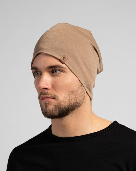 A studio portrait of a man looking thoughtfully off to the left side of the frame. He is wearing a light beige, slouchy knit beanie featuring a small, square brown leather logo patch on the front-left edge. He is also wearing a solid black crew-neck top. The background is a plain light grey.