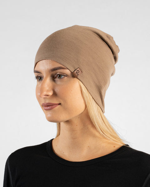 A studio portrait of a blonde woman looking thoughtfully off to the left side of the frame. She is wearing a light beige, slouchy knit beanie featuring a small, square brown leather logo patch on the front-left edge. She is also wearing a solid black long-sleeved top. The background is a plain light grey.