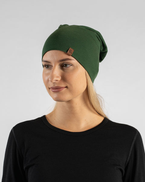 A studio portrait of a blonde woman looking off to the left against a plain light grey background. She is wearing a black long-sleeved shirt and a dark green slouchy beanie featuring a small brown logo tag.
