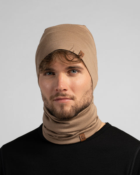 A studio portrait of a man looking directly at the camera against a plain light grey background. He is wearing a black shirt paired with a matching beige beanie and neck gaiter.