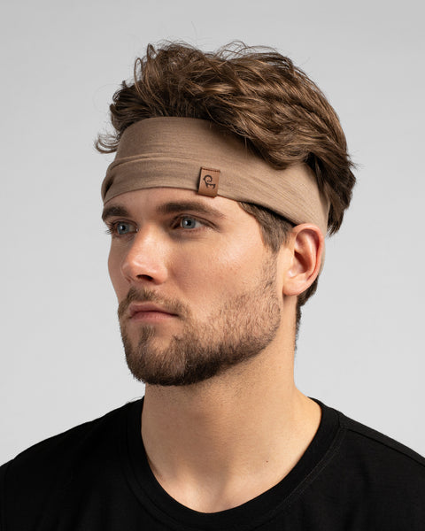 A close-up studio portrait of a man looking thoughtfully off to the left side of the frame. He is wearing a wide, light beige knit headband that pushes back his wavy brown hair, paired with a black sleeveless top. A small, square brown leather logo patch is featured on the front-left side of the headband. The background is a solid light grey.