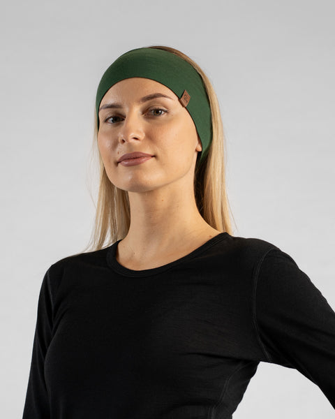 A studio portrait of a blonde woman looking directly at the camera with her body slightly angled. She is wearing a black long-sleeved shirt and a wide dark green headband with a small brown logo tag against a light grey background.