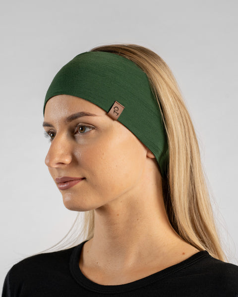 A studio portrait of a blonde woman shown in a three-quarter profile looking off to the left against a plain light grey background. She is wearing a black long-sleeved shirt and a dark green headband featuring a small brown logo tag.