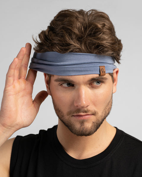 A studio portrait of a man looking off to the right, wearing a dusty blue headband and a black sleeveless shirt. He is raising his right hand as if adjusting the headband.