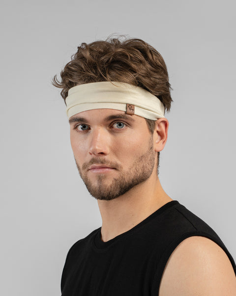 A studio portrait of a man looking directly at the camera. He has wavy brown hair and a short beard. He is wearing a light cream, natural-colored headband featuring a small brown leather logo patch on the front-left edge. He is wearing a black sleeveless top against a plain light grey background.