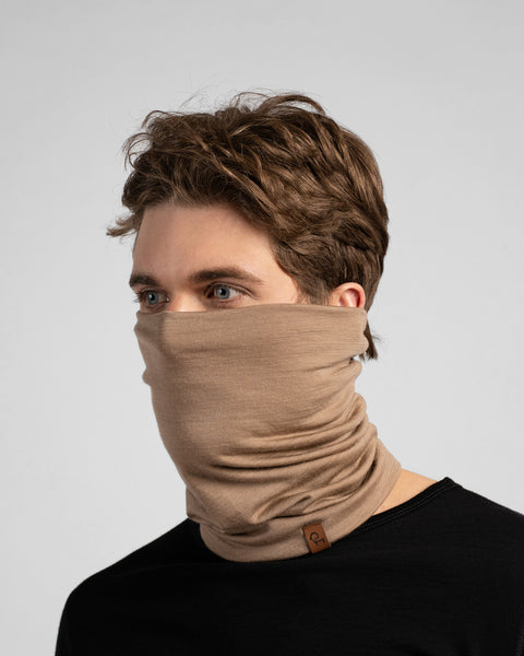 A studio portrait of a man looking thoughtfully off to the left side of the frame. He is wearing a light beige neck gaiter pulled up over his nose, covering his lower face. The gaiter has a small brown leather logo patch on the bottom edge. He is wearing a solid black long-sleeved top against a plain light grey background.