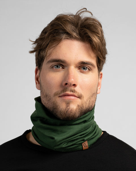 A studio portrait of a man looking directly at the camera against a light grey background. He is wearing a black long-sleeved shirt and a dark green neck gaiter bunched around his neck, featuring a small brown logo tag.