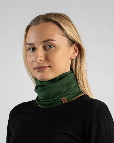A studio portrait of a blonde woman looking directly at the camera. She is wearing a black long-sleeved shirt and a dark green neck gaiter bunched loosely around her neck, featuring a small brown logo tag against a light grey background.