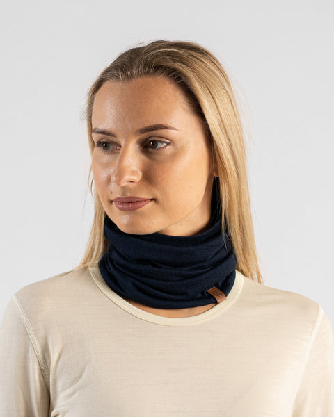 A studio portrait of a blonde woman looking slightly off to the left against a light grey background. She is wearing a light beige long-sleeved shirt and a navy blue neck gaiter bunched loosely around her neck, featuring a small brown logo tag at the bottom edge.