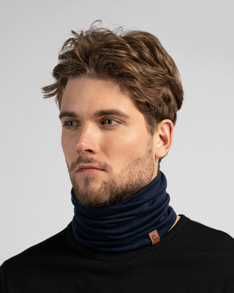 A studio portrait of a man looking off to the left, wearing a black long-sleeved shirt and a navy blue neck gaiter. The gaiter is scrunched around his neck and features a small brown logo tag.