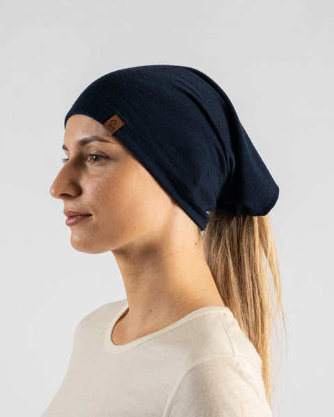 A side profile studio portrait of a blonde woman facing left against a plain light grey background. She is wearing a light beige long-sleeved shirt and a navy blue neck gaiter worn over her head as a hair wrap, with her ponytail extending out the back. A small brown logo tag is visible on the side of the gaiter.