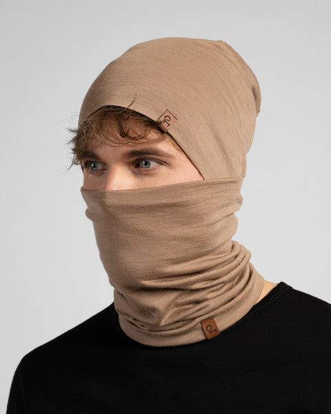 A studio portrait of a man looking thoughtfully off to the left side of the frame. He is wearing both a light beige slouchy beanie and a matching light beige neck gaiter pulled up to cover his nose and lower face. Both items feature a small brown leather logo patch. He is wearing a solid black long-sleeved top against a plain light grey background.