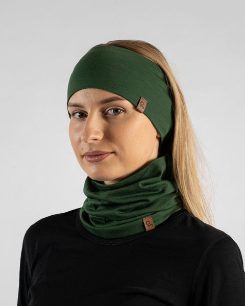 A studio portrait of a blonde woman standing at a slight angle, looking towards the camera against a plain light grey background. She is wearing a black long-sleeved shirt and a matching dark green set consisting of a wide headband and a neck gaiter pulled up to cover her lower face. Both accessories feature small brown logo tags.