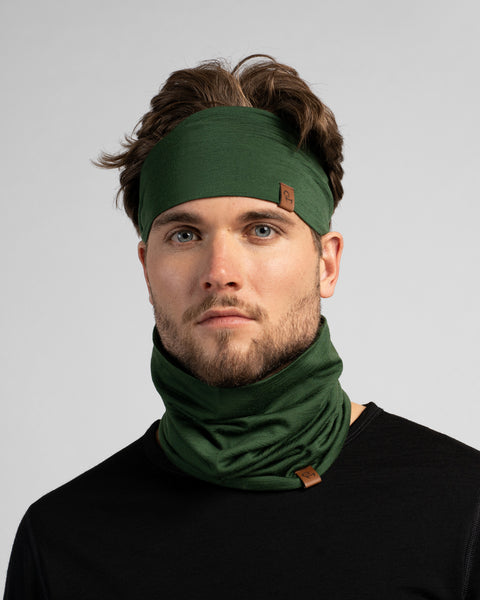 A studio portrait of a man looking directly at the camera against a light grey background. He wears a black shirt, a dark green headband holding back his wavy brown hair, and a matching dark green neck gaiter bunched around his neck.